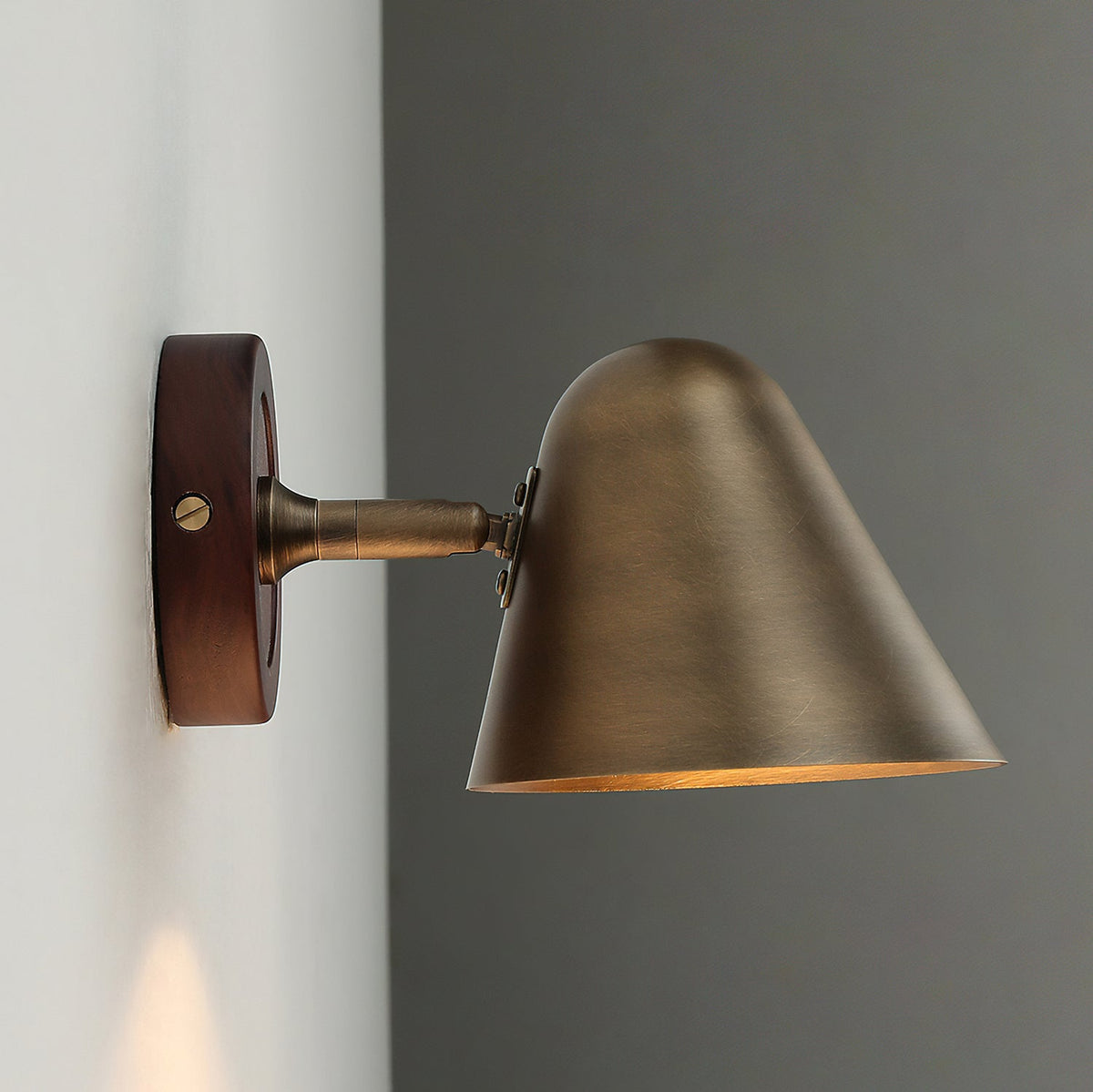 Profile view of a Clérachic antique brass wall light on a dark walnut-coloured backplate, standing out from a wall corner.