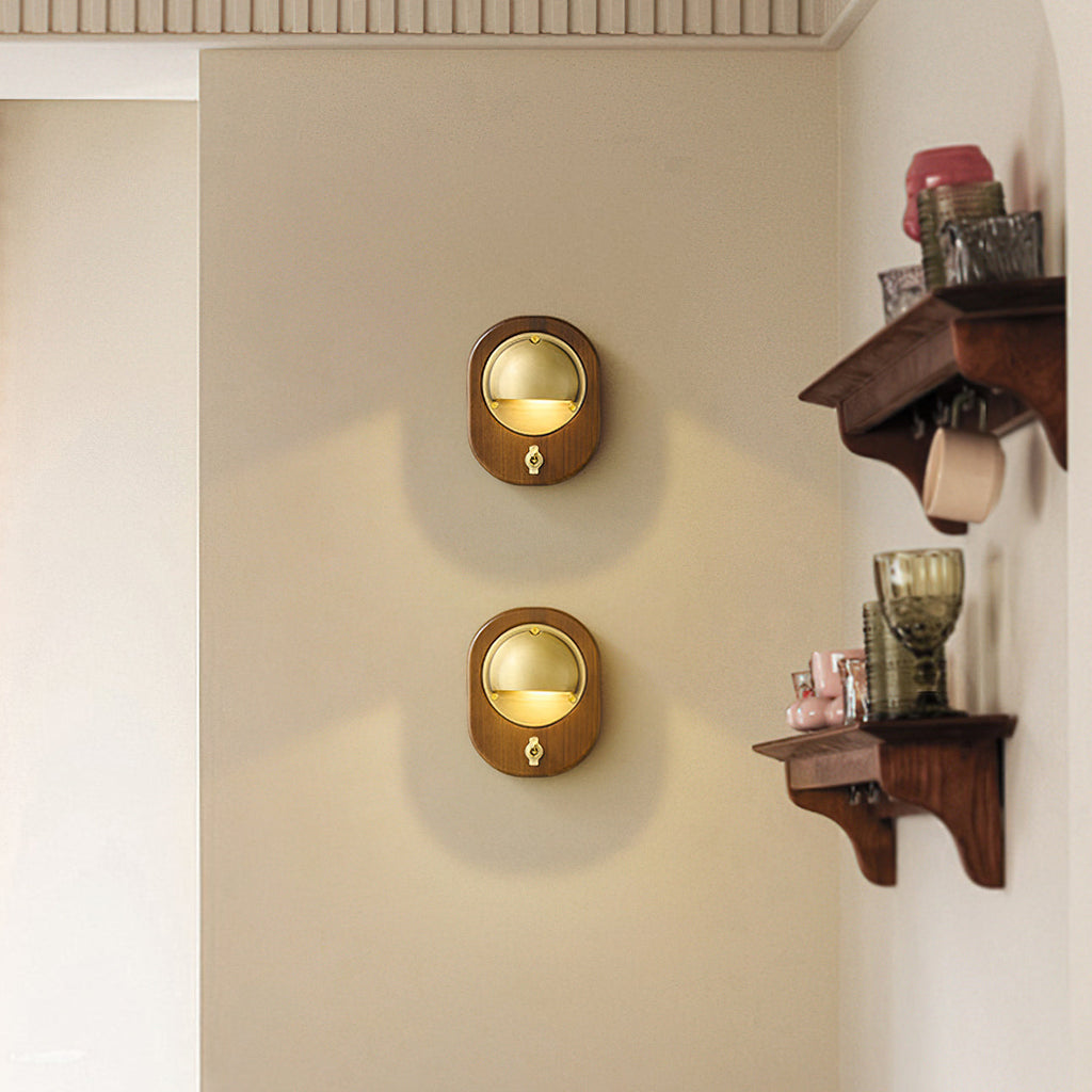 Two oval Walnut & Brass Clérachic wall lights stacked on a cream wall, beside a dark wooden shelf with glassware and cups.