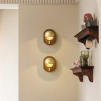 Two oval Walnut & Brass Clérachic wall lights stacked on a cream wall, beside a dark wooden shelf with glassware and cups.