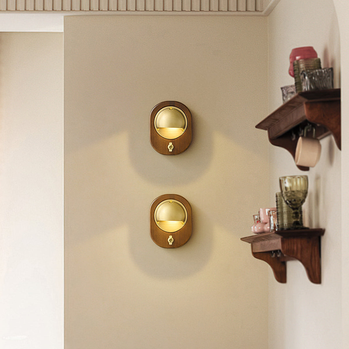 Two oval Walnut & Brass Clérachic wall lights stacked on a cream wall, beside a dark wooden shelf with glassware and cups.