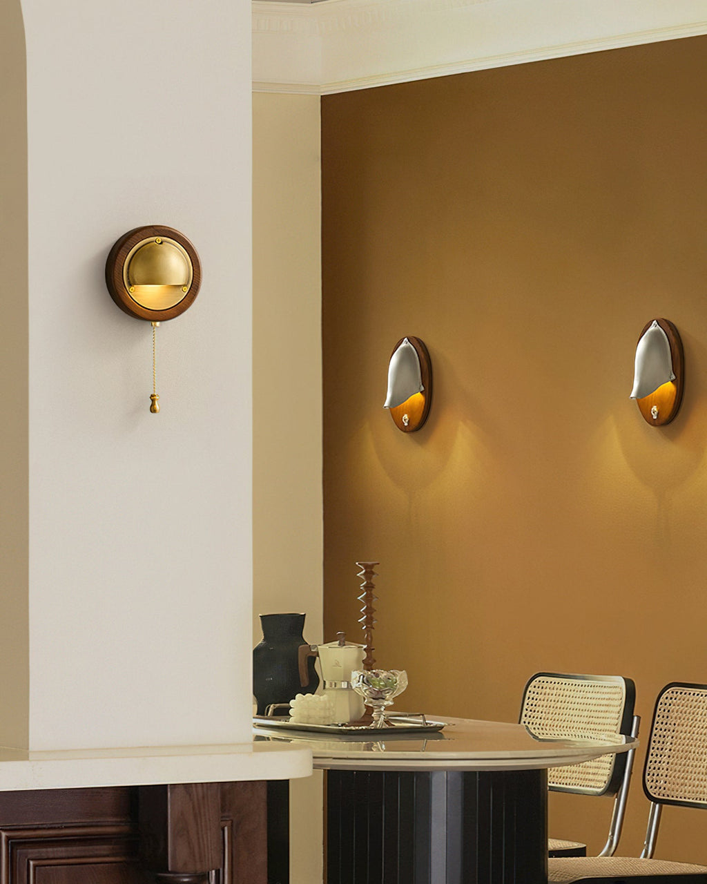 A round Clérachic wall light with a pull chain on a cream wall, with an amber-painted dining corner and two more lights beyond.