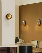 A round Clérachic wall light with a pull chain on a cream wall, with an amber-painted dining corner and two more lights beyond.