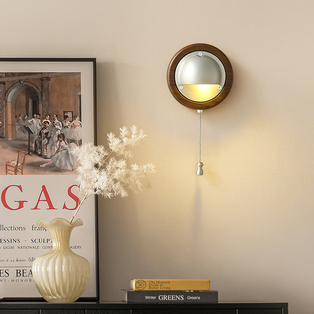 A Walnut & Silver Clérachic wall light with a pull chain glows beside a framed print and a vase of dried flowers on a dark sideboard.