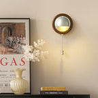 A Walnut & Silver Clérachic wall light with a pull chain glows beside a framed print and a vase of dried flowers on a dark sideboard.