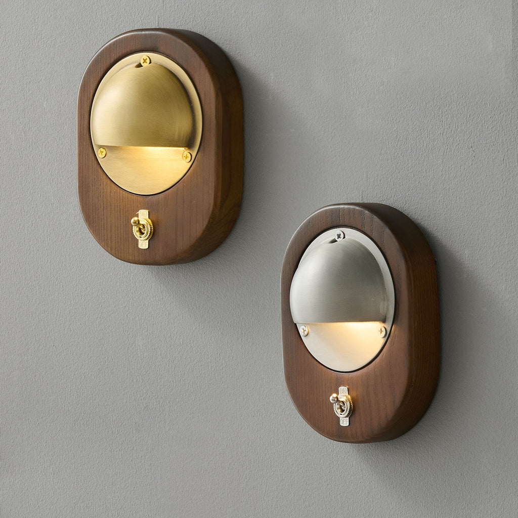 An oval Walnut & Brass and an oval Walnut & Silver Clérachic wall light side by side on a grey wall, each with a small toggle switch.