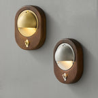 An oval Walnut & Brass and an oval Walnut & Silver Clérachic wall light side by side on a grey wall, each with a small toggle switch.