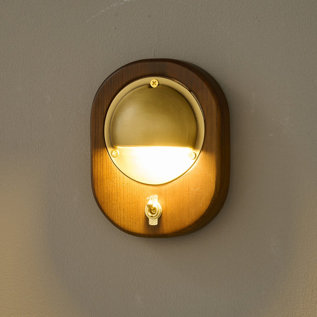 A lit oval Walnut & Brass Clérachic wall light with a small brass toggle switch, glowing warm against a dark wall.