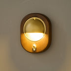A lit oval Walnut & Brass Clérachic wall light with a small brass toggle switch, glowing warm against a dark wall.