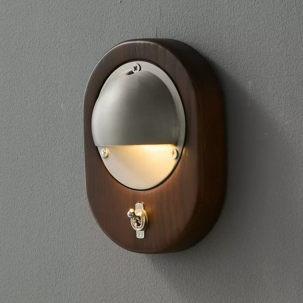 Seen slightly from the side, an oval Walnut & Silver Clérachic wall light shows its deep walnut frame and silver toggle switch.