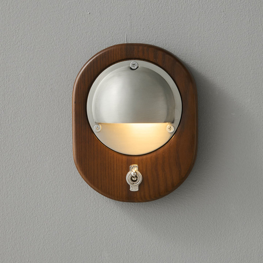 An oval Walnut & Silver Clérachic wall light on a grey wall, with warm light washing down from under the half-dome shade.
