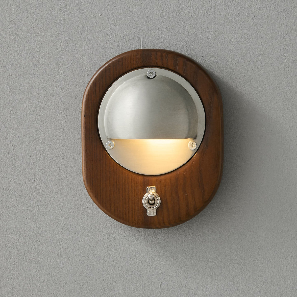 An oval Walnut & Silver Clérachic wall light on a grey wall, with warm light washing down from under the half-dome shade.