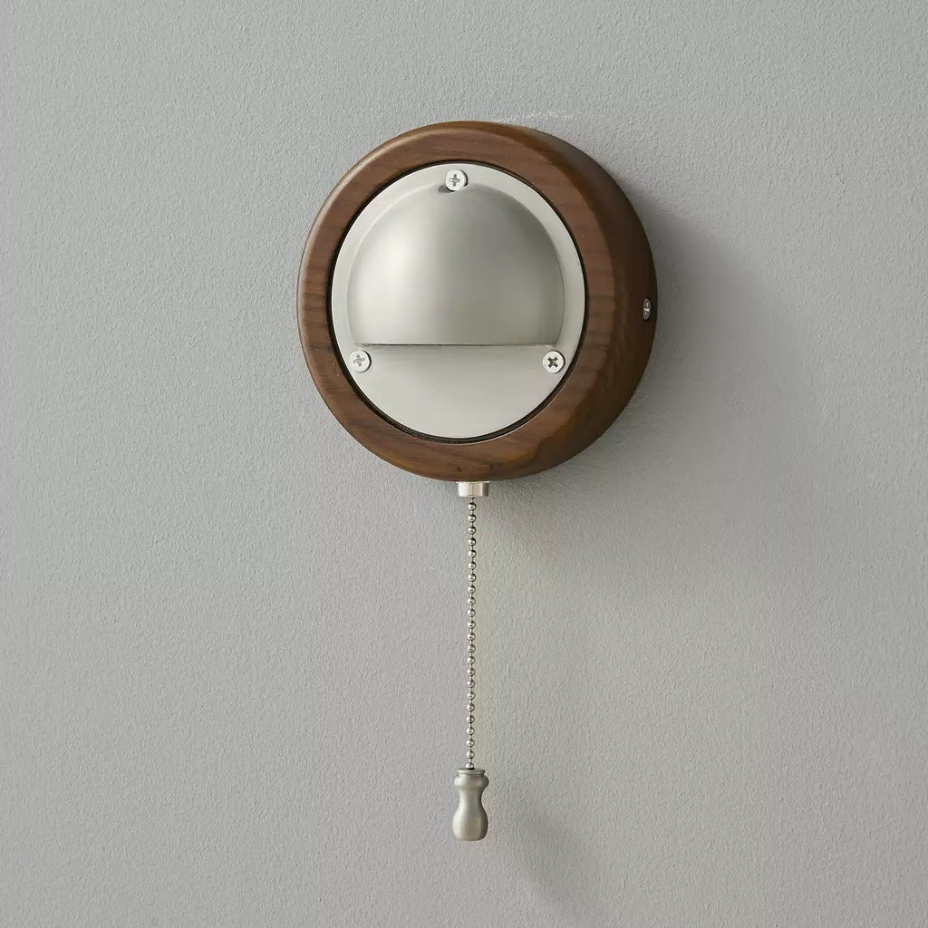 A small round Walnut & Silver Clérachic wall light with its beaded pull chain and silver knob hanging on a grey wall.