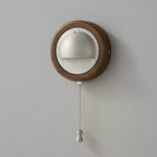 A small round Walnut & Silver Clérachic wall light with its beaded pull chain and silver knob hanging on a grey wall.