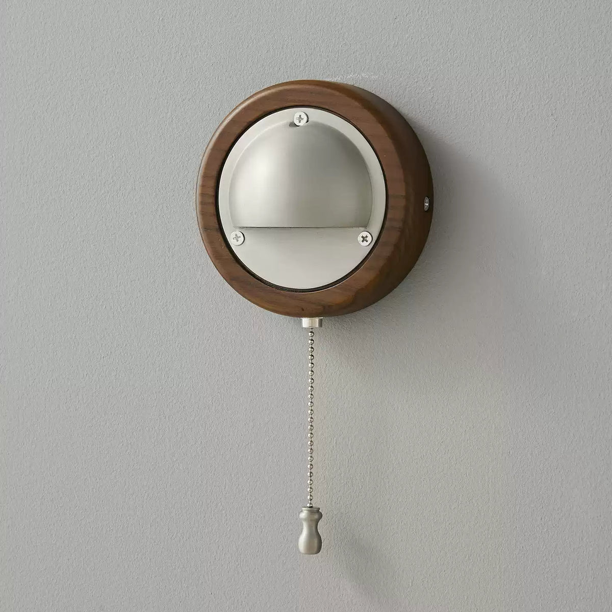 A small round Walnut & Silver Clérachic wall light with its beaded pull chain and silver knob hanging on a grey wall.
