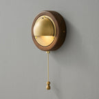 A round Walnut & Brass Clérachic wall light with a lit brass half-dome and a brass pull chain and knob, on a grey wall.