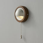 A round Walnut & Silver Clérachic wall light, lit, with its fine chain hanging below on a grey wall.