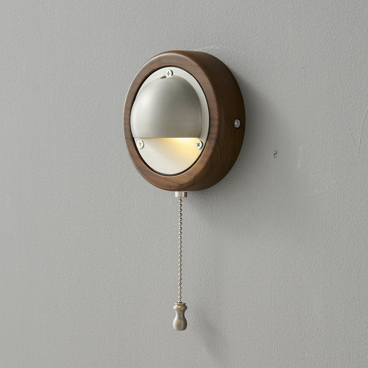A round Walnut & Silver Clérachic wall light, lit, with its fine chain hanging below on a grey wall.