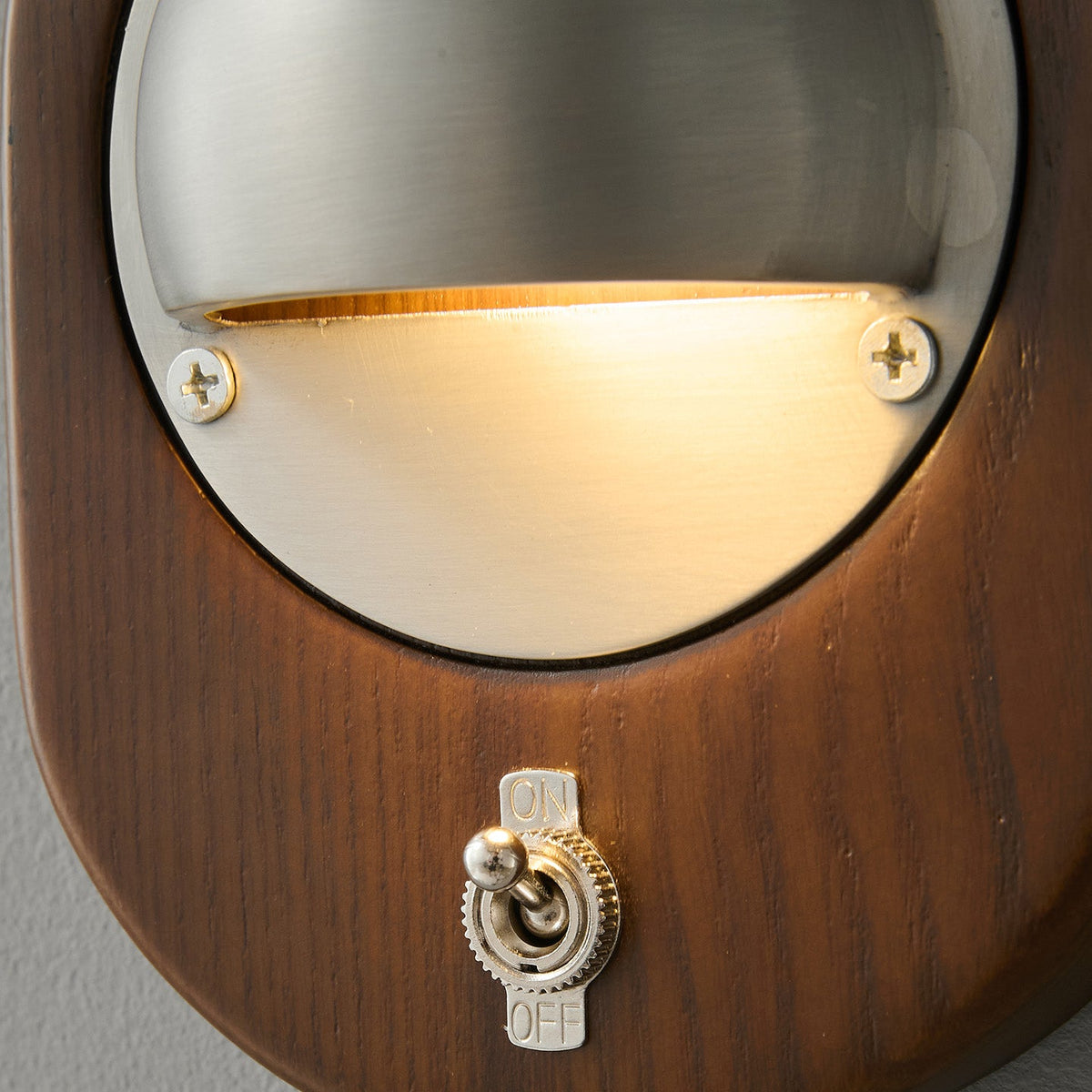 Close view of the lit silver half-dome, the walnut grain and the On/Off toggle switch on this Clérachic oval wall light.