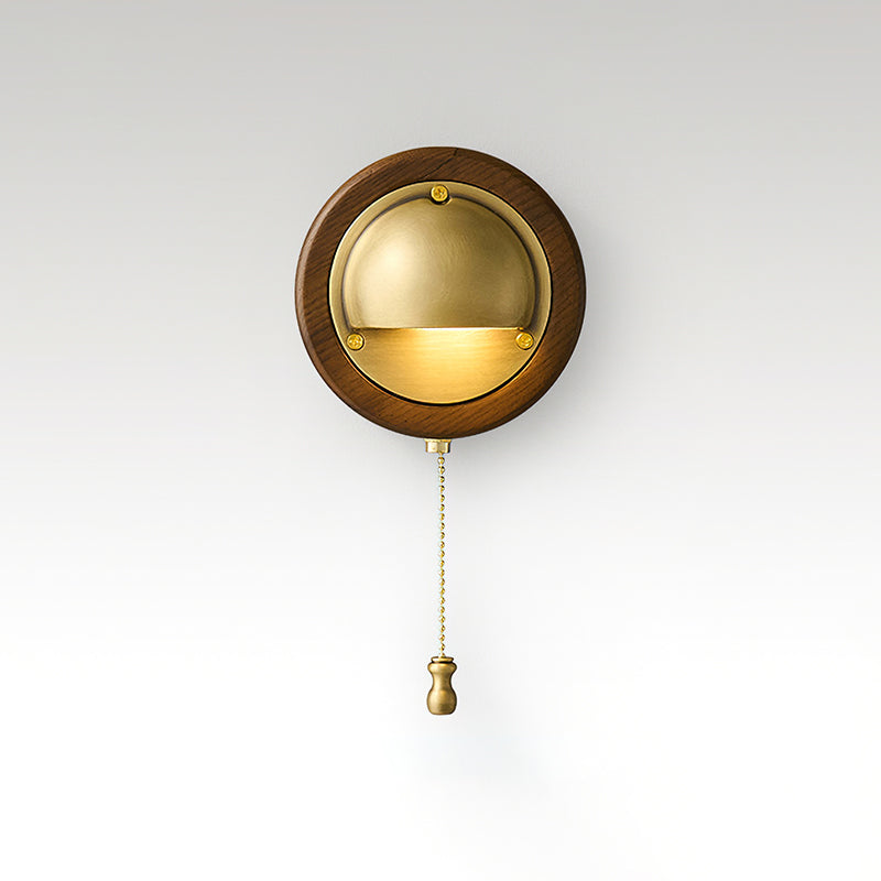 Round Walnut & Brass Clérachic wall light with a lit half-dome and a brass chain pull, on a soft grey studio backdrop.