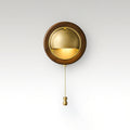 Round Walnut & Brass Clérachic wall light with a lit half-dome and a brass chain pull, on a soft grey studio backdrop.
