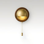 Round Walnut & Brass Clérachic wall light with a lit half-dome and a brass chain pull, on a soft grey studio backdrop.