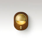 Oval Walnut & Brass Clérachic wall light with a brass toggle switch below the lit half-dome, on a soft grey studio backdrop.
