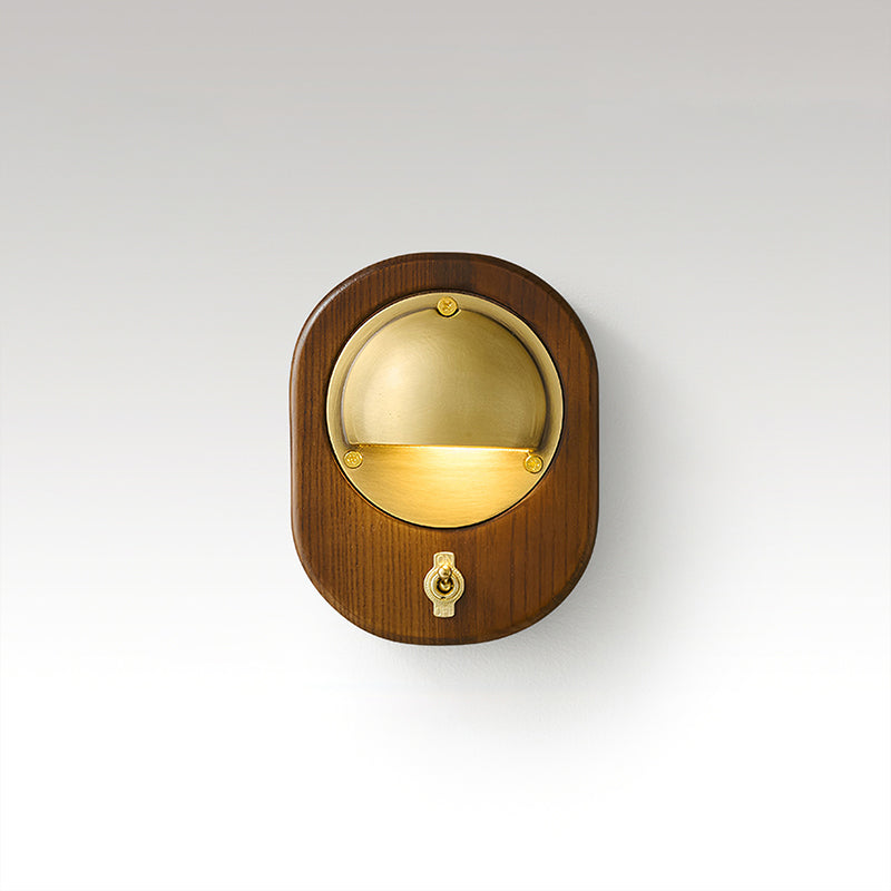 Oval Walnut & Brass Clérachic wall light with a brass toggle switch below the lit half-dome, on a soft grey studio backdrop.