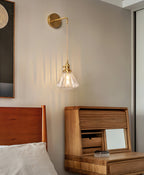 A Clérachic wall light hangs above a wooden bedside cabinet with a roll-top front, beside a bed with a wooden headboard.