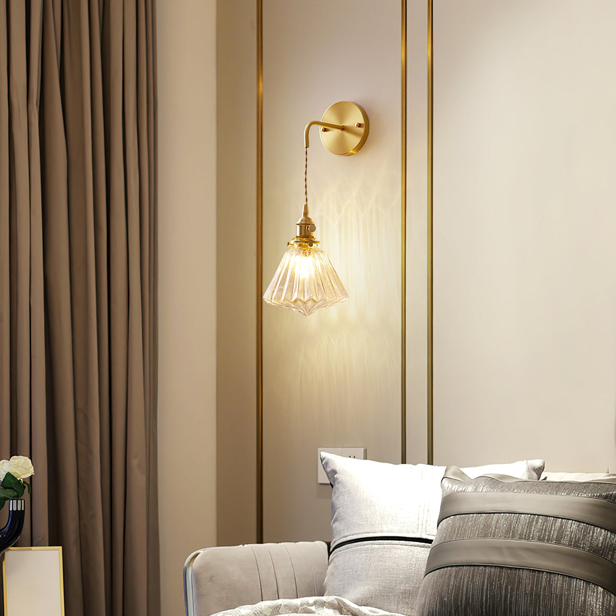 A Clérachic wall light lit beside a brown curtain and a bed with grey cushions, on a wall with slim brass trim.