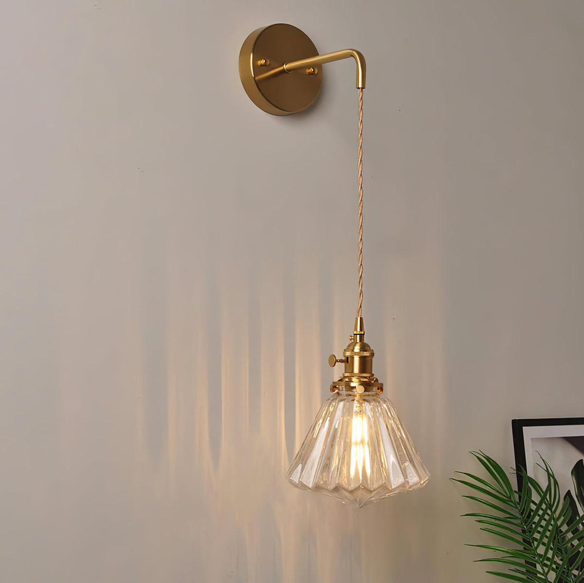 A Clérachic wall light hangs on a grey-beige wall beside a framed picture and the tips of a green palm leaf.