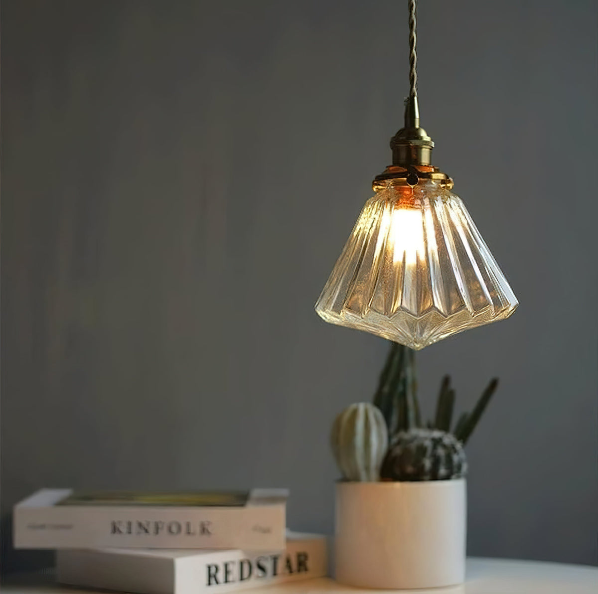 A Clérachic pendant-style light with a ribbed glass shade glows against a dark wall, above a cactus pot and a stack of books.