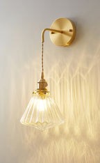 A Clérachic wall light with a round brass backplate and a lit, ribbed glass shade, its light patterned on a grey wall.