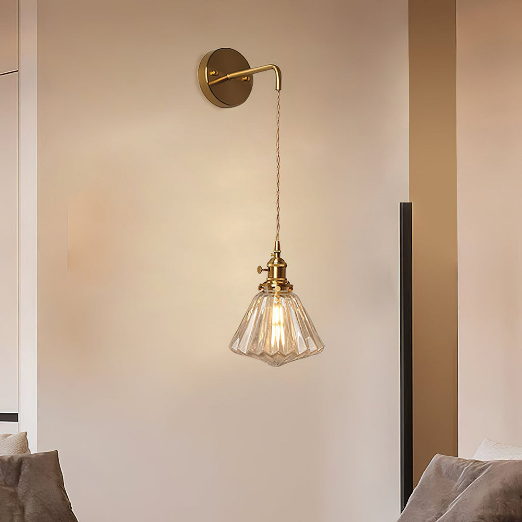 A Clérachic wall light on a pale wall above a sofa, its slim brass arm holding a small ribbed glass shade on a long cord.