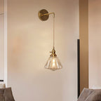 A Clérachic wall light on a pale wall above a sofa, its slim brass arm holding a small ribbed glass shade on a long cord.