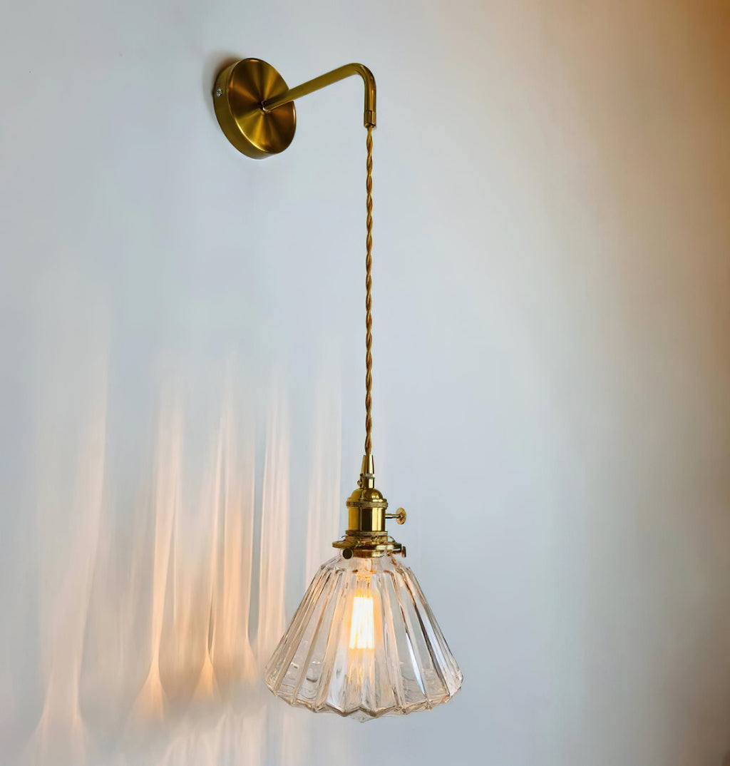 A Clérachic wall light hangs on a pale blue-grey wall, its lit glass shade casting streaks of warm light behind.