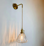 A Clérachic wall light hangs on a pale blue-grey wall, its lit glass shade casting streaks of warm light behind.