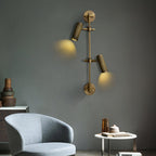 A Clérachic wall light with two aimed spot heads throws pools of warm light down a grey-green wall, beside a pale blue armchair and side table.