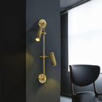 Two-head Clérachic wall light on a dark wall beside a window, with a pool of warm light below each head and a black chair nearby.