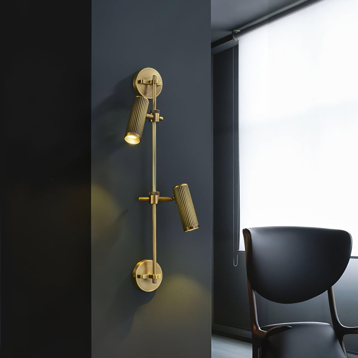 Two-head Clérachic wall light on a dark wall beside a window, with a pool of warm light below each head and a black chair nearby.
