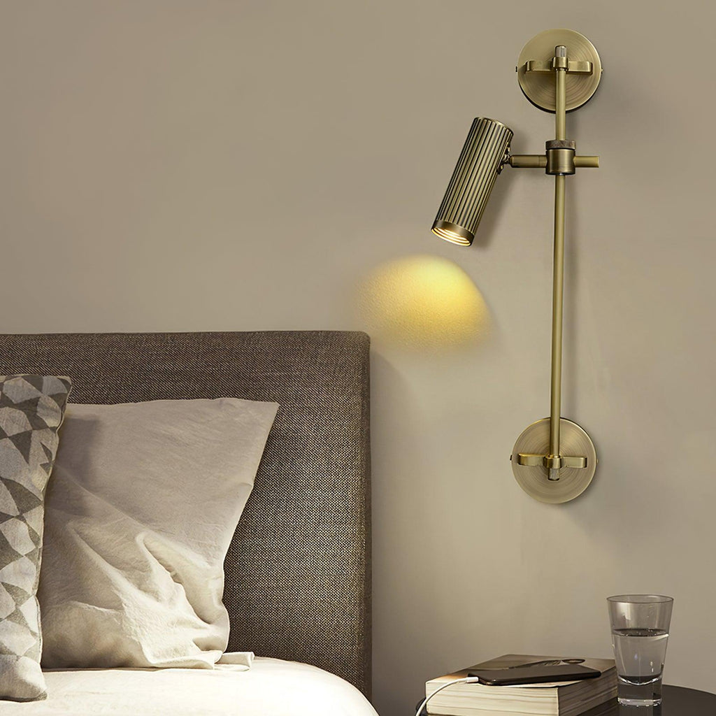 Single-head Clérachic wall light over a bedside, with the fluted bronze cylinder aimed down at a grey headboard, a book and a glass of water.
