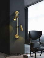 Two bronze spot heads, lit, from Clérachic on a dark wall beside a window, with a black armchair and stacked books beneath.
