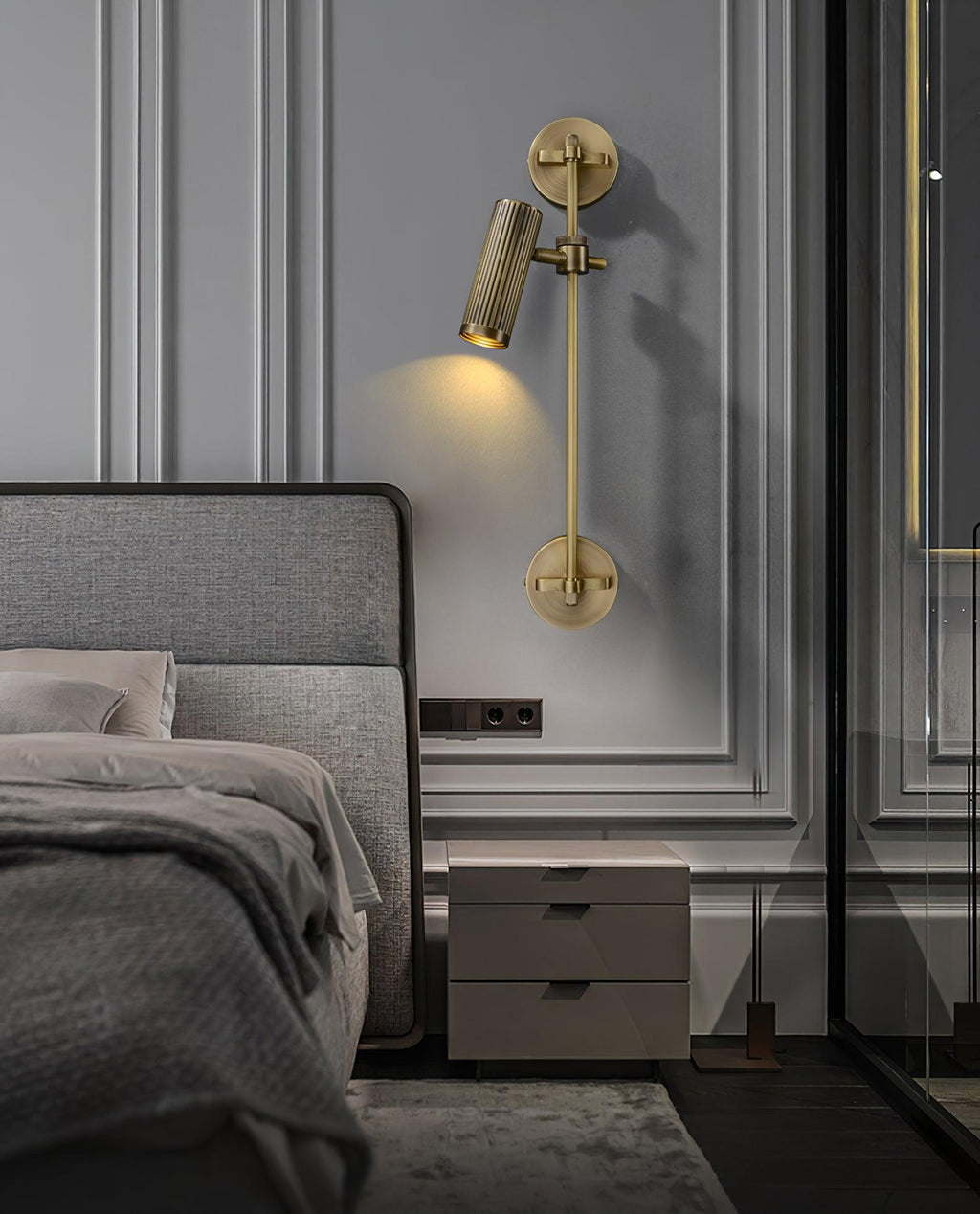 A Clérachic wall light with a single head aimed down, mounted on panelled grey wall beside a double bed and a pale bedside cabinet.