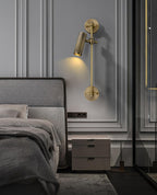 A Clérachic wall light with a single head aimed down, mounted on panelled grey wall beside a double bed and a pale bedside cabinet.