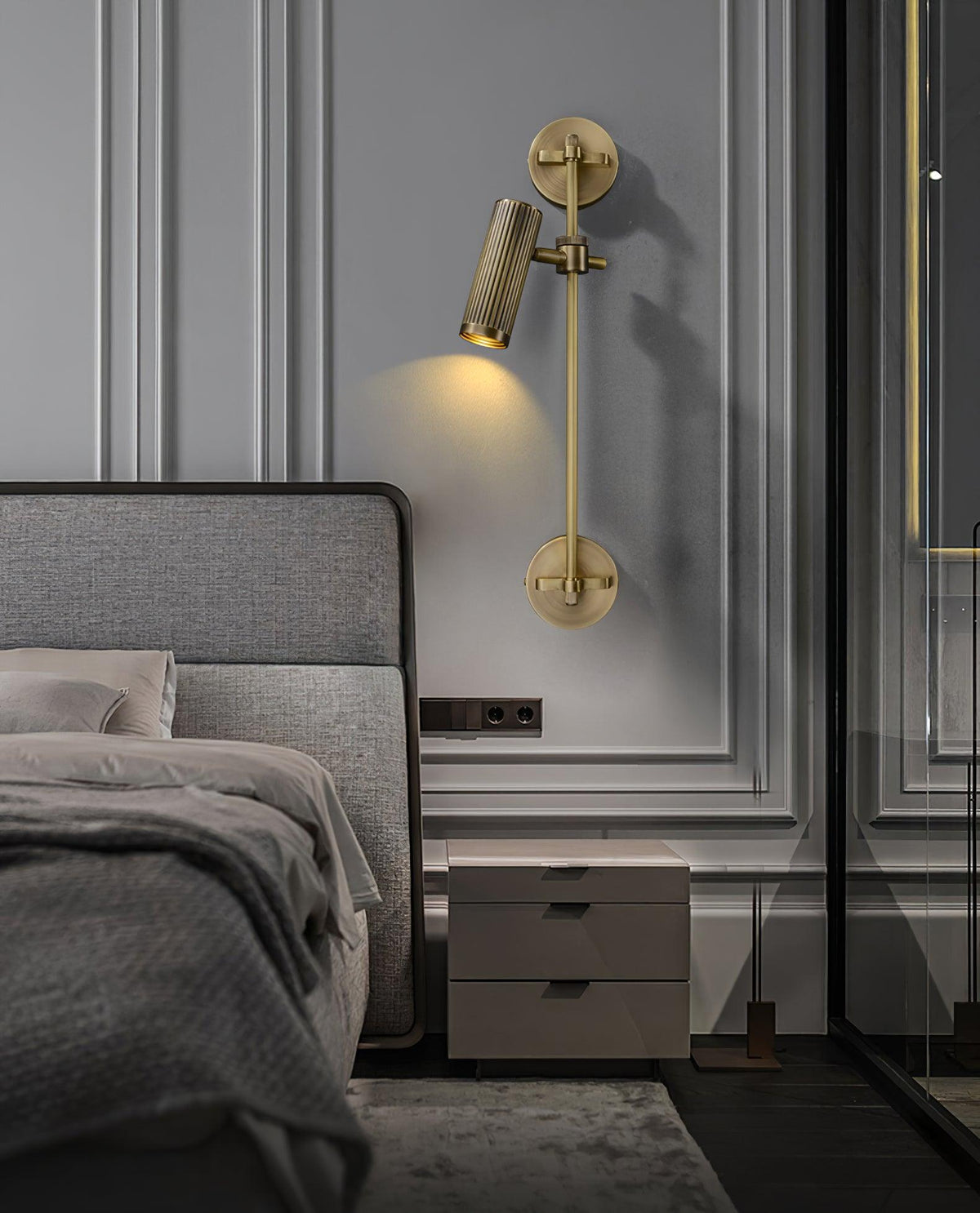 A Clérachic wall light with a single head aimed down, mounted on panelled grey wall beside a double bed and a pale bedside cabinet.