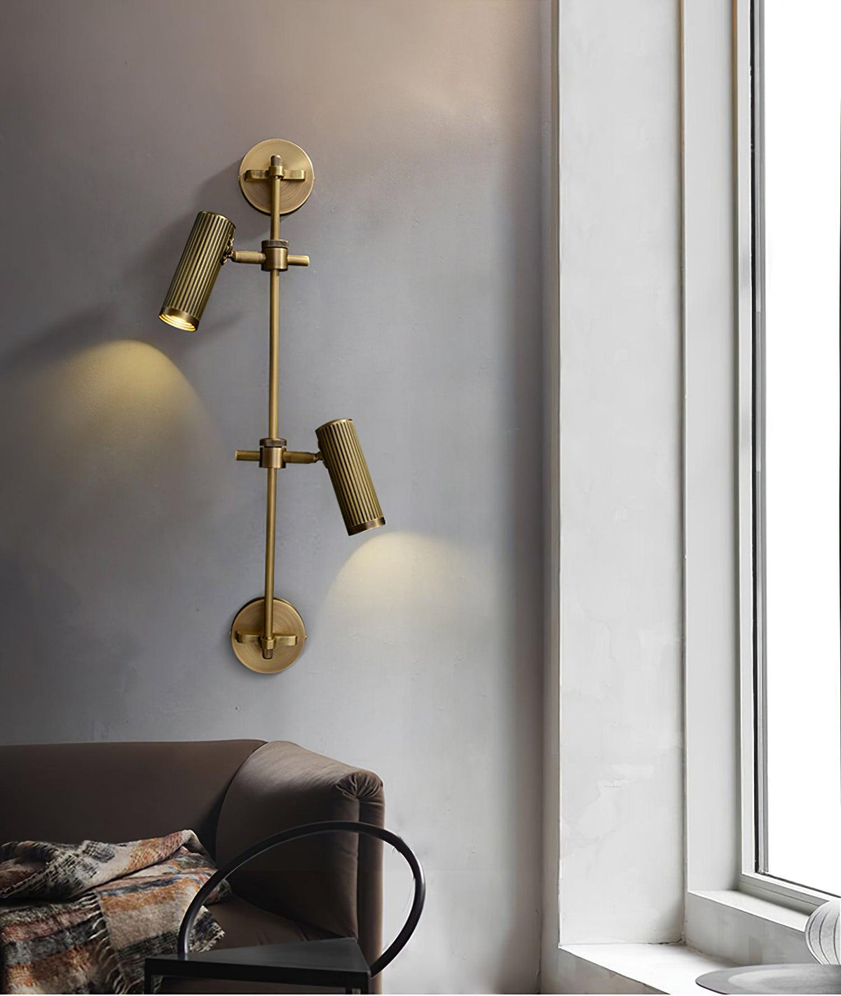 Two-head Clérachic wall light in bronze, lit, beside a window with a brown sofa and a patterned throw in the lower corner.