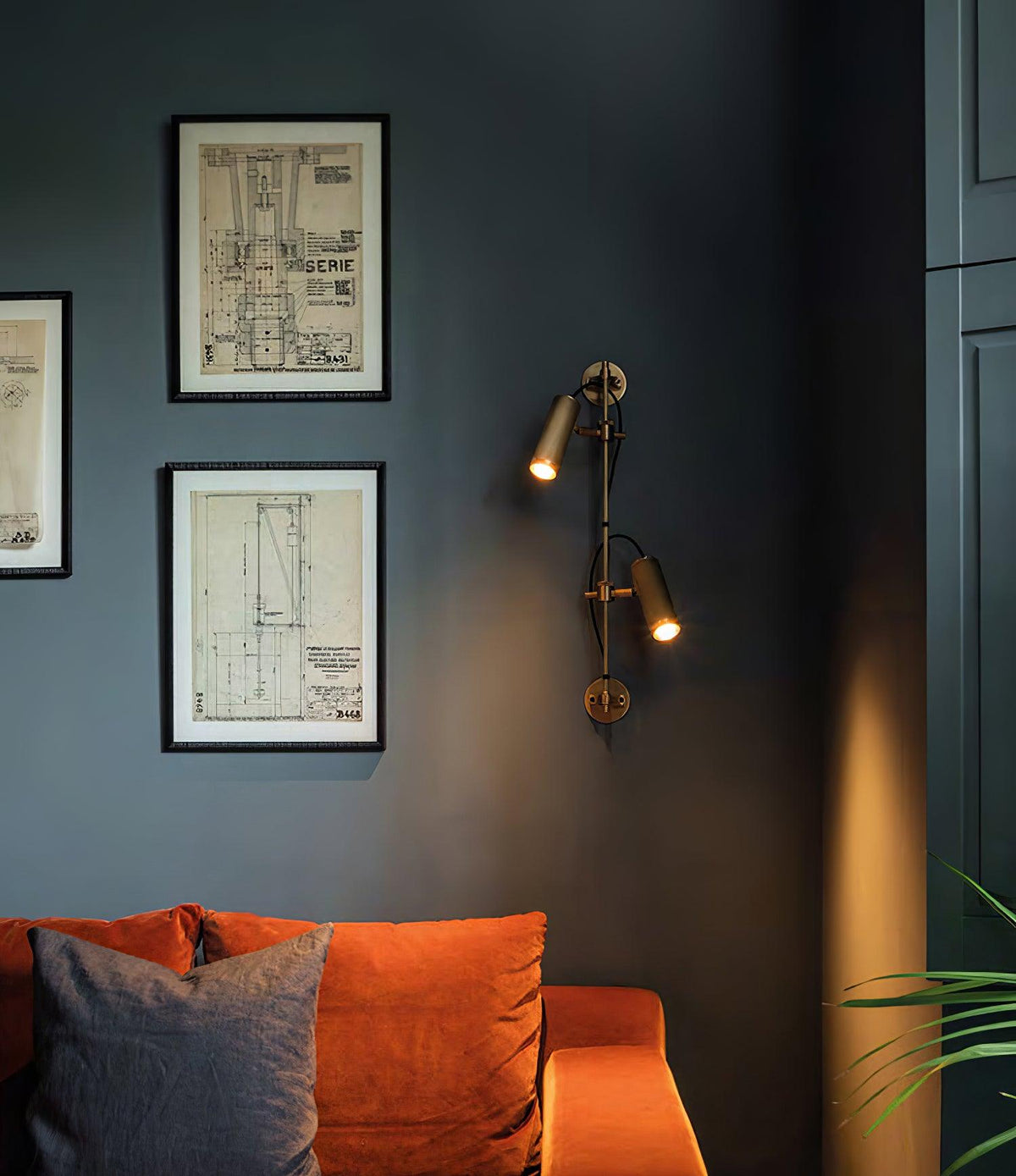 Two bronze heads on a slim rail from Clérachic, lit, on a dark blue-grey wall with framed technical drawings and an orange sofa.