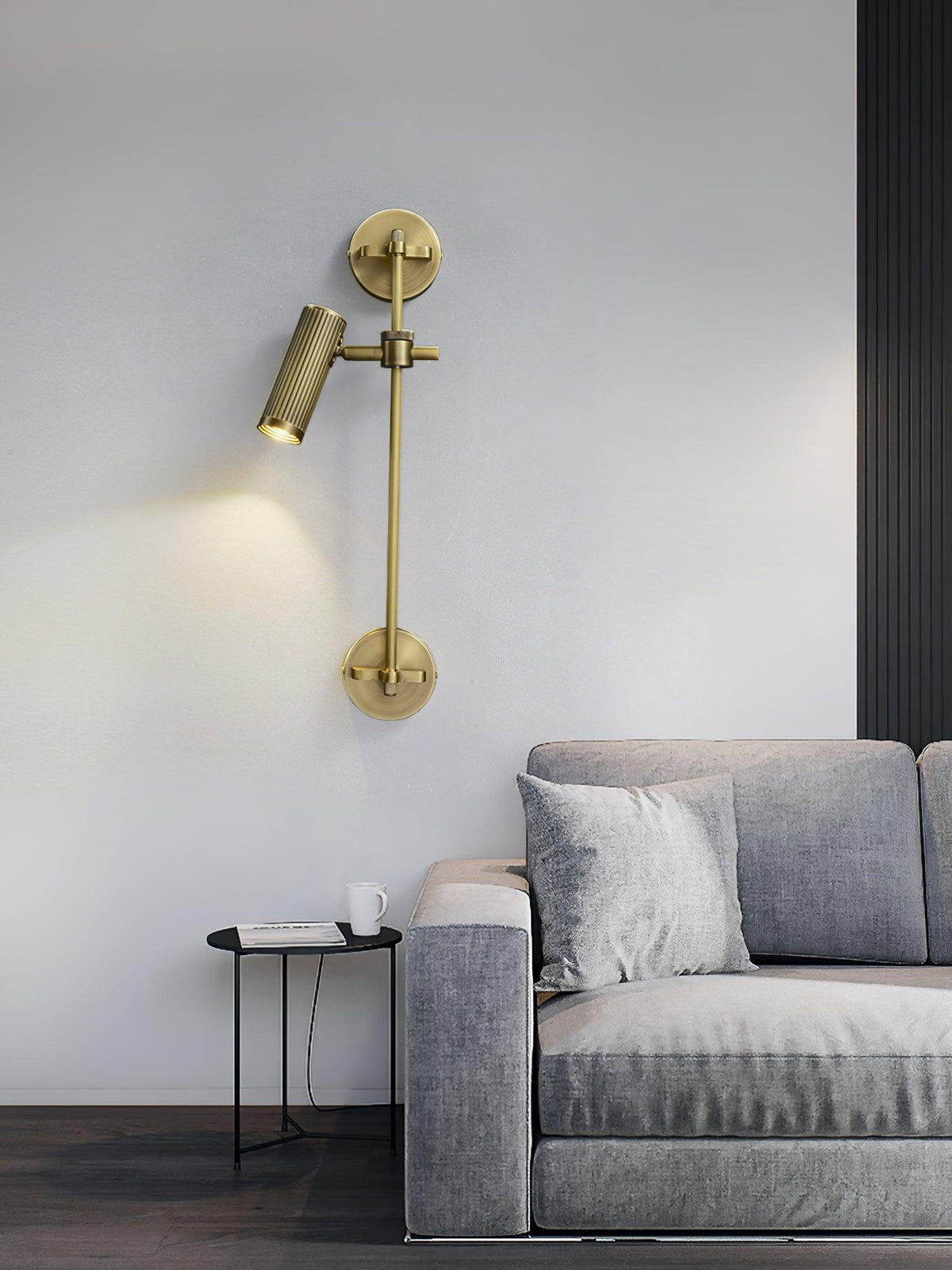 A single-head bronze wall light from Clérachic, lit, on a pale wall above a grey sofa, with a black side table and a cup beside it.