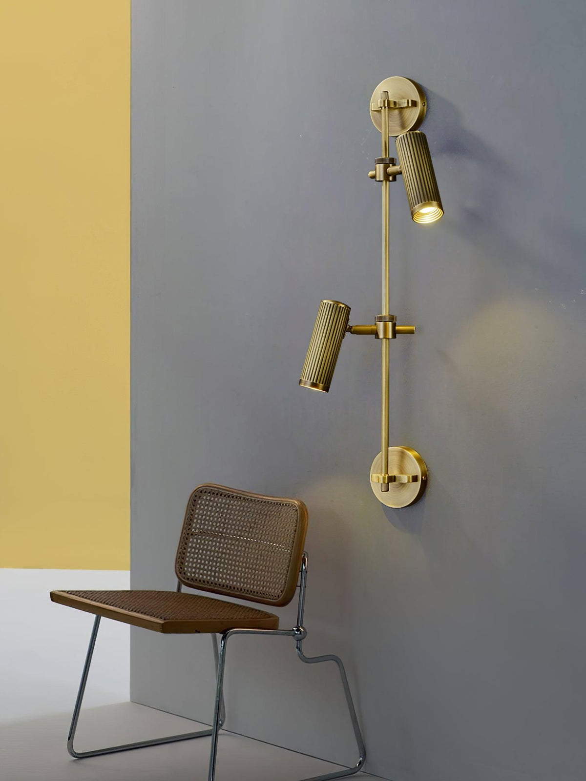 Two-head wall light from Clérachic on a grey-blue wall with a yellow panel, with each head aimed down above a cane-backed chair.