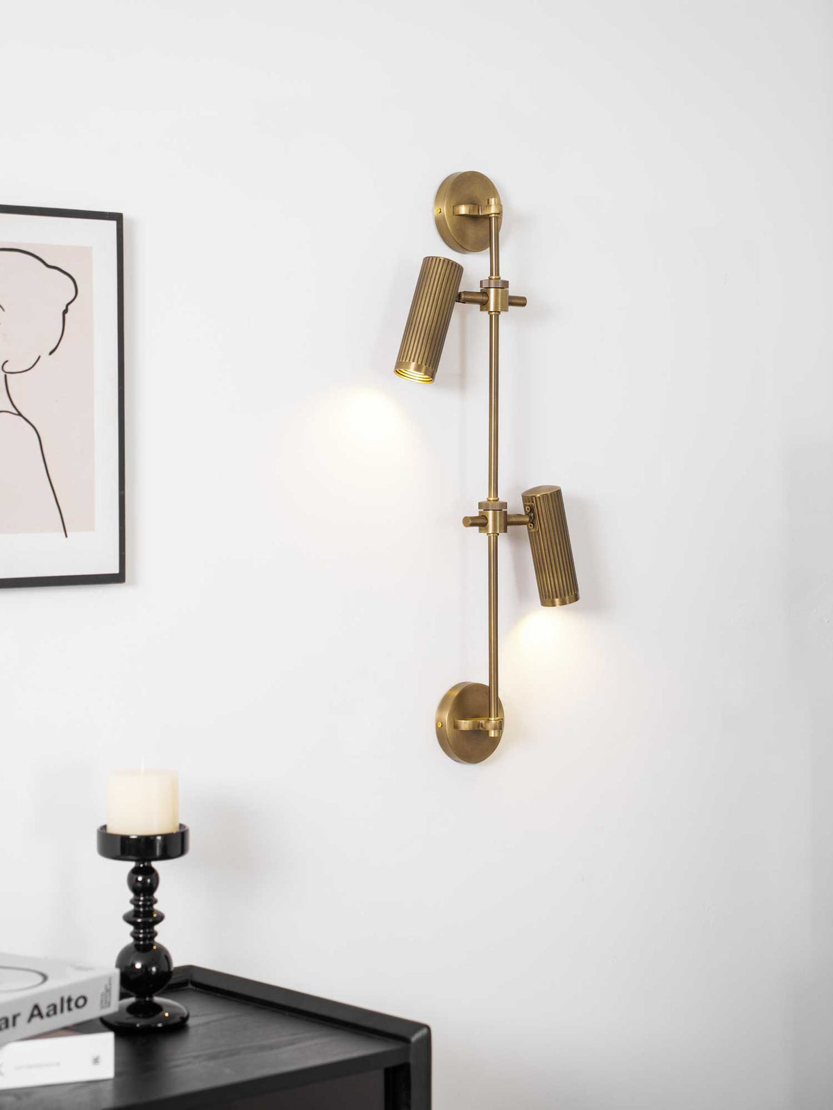 Two bronze spot heads from Clérachic on a white wall, lit, with a framed line drawing and a black candlestick on a dark side table.