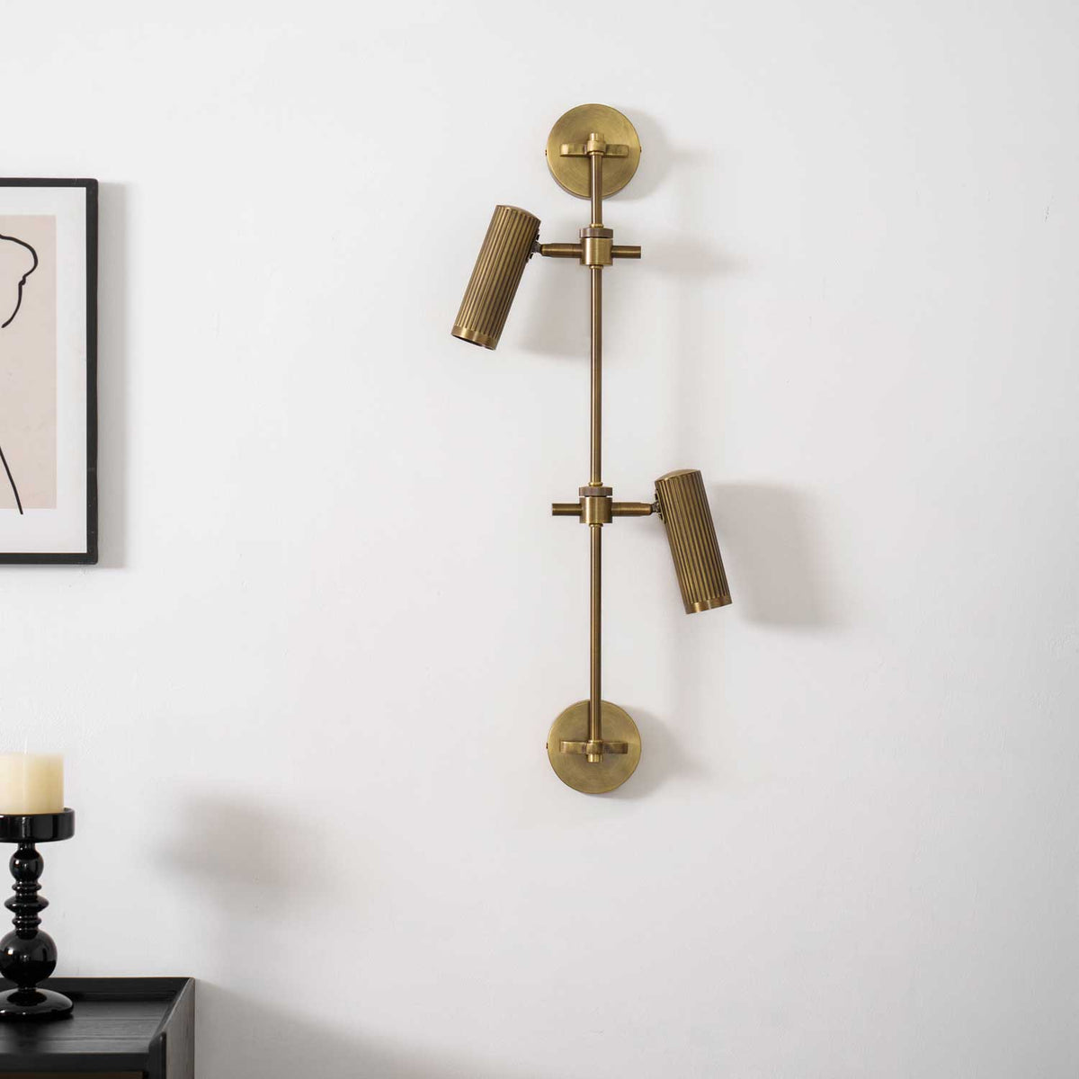 Two fluted bronze heads on a vertical rail from Clérachic, close to a white wall with a framed print and a candlestick below.